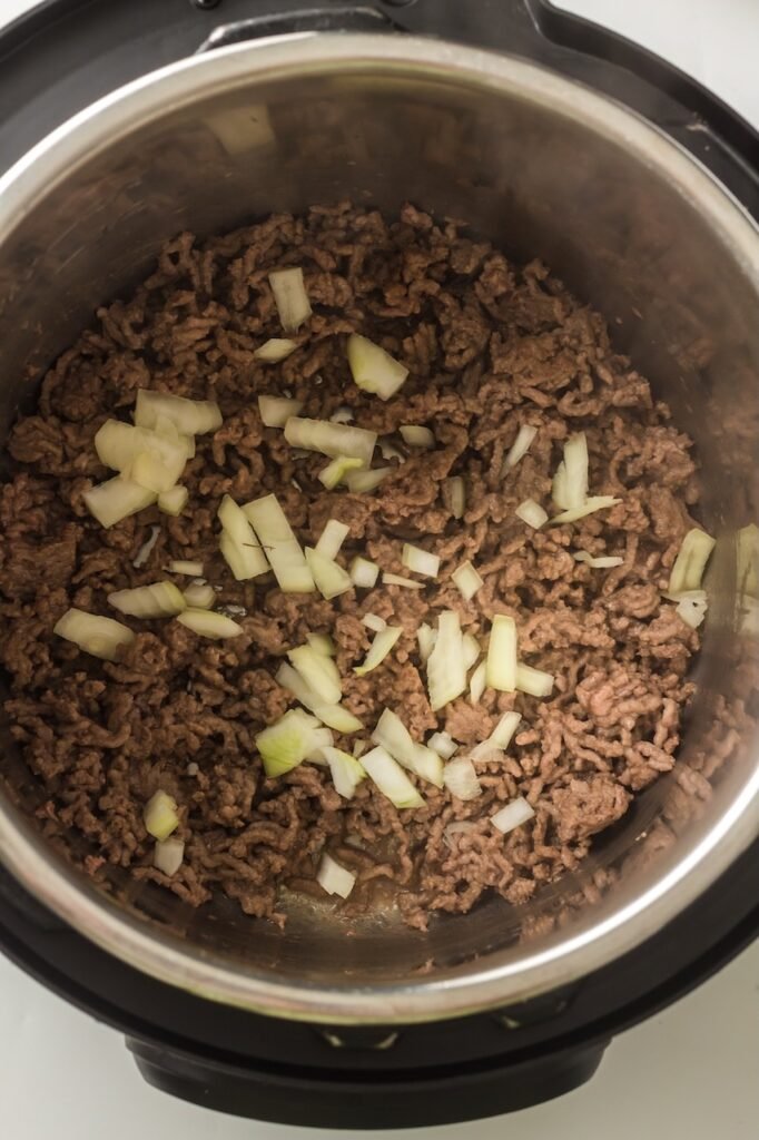 Diced onion scattered over fully browned ground beef inside  insert, showing the onions just added to the cooked meat during the sauté step before spices are mixed in.