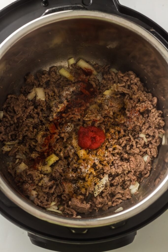 Tomato paste and dry spices added to browned ground beef and onions inside insert, showing the seasonings sitting on top of the meat before being stirred and cooked during the sauté step.