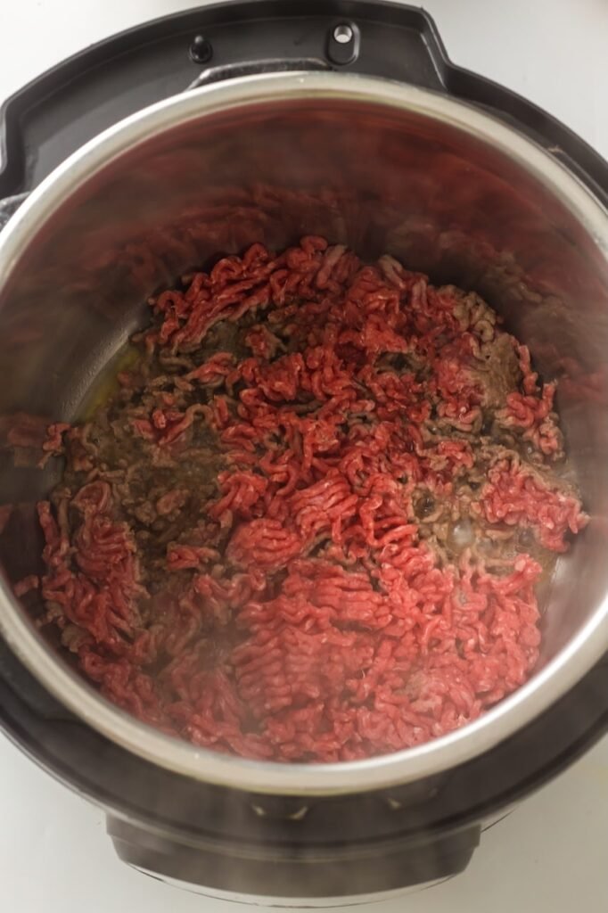 Ground beef browning inside an Instant Pot insert, showing partially cooked meat breaking apart as it cooks, with steam rising and the beef beginning to lose its pink color during the sauté step.
