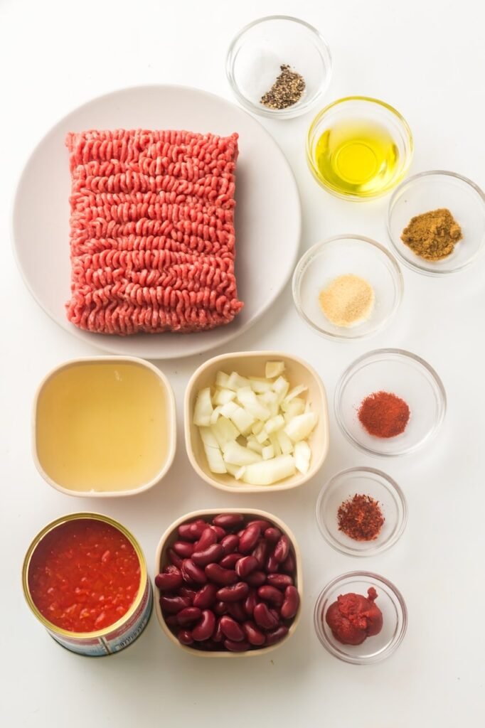 A top-down view of the ingredients for Instant Pot chili, including raw ground beef on a plate, diced onion, kidney beans, diced tomatoes, beef broth, olive oil, tomato paste, and small bowls of chili powder, cumin, paprika, garlic powder, salt, and black pepper arranged on a white surface.