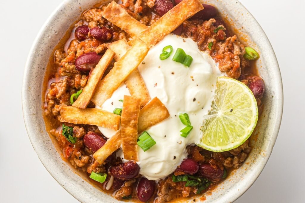 An overhead view of a bowl of Instant Pot chili made with ground beef and kidney beans, topped with sour cream, chopped green onions, crispy tortilla strips, and a lime wedge, showing the thick texture and rich color of the chili.