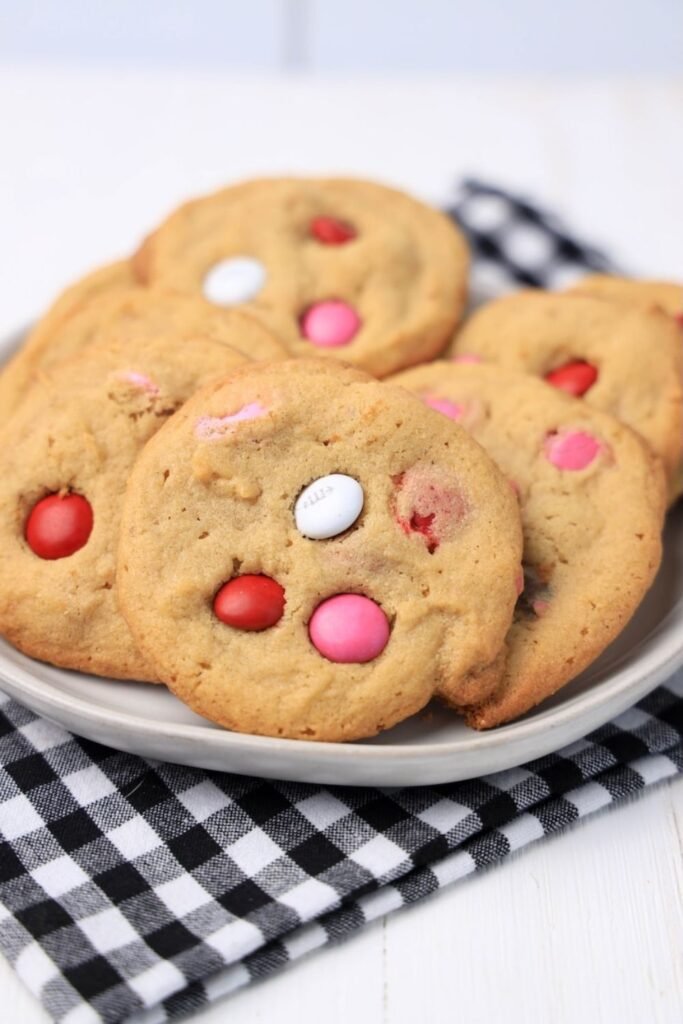 A plate of soft Valentine’s M&M cookies stacked on a black-and-white cloth, showing golden peanut butter cookies studded with red, pink, and white M&M candies.