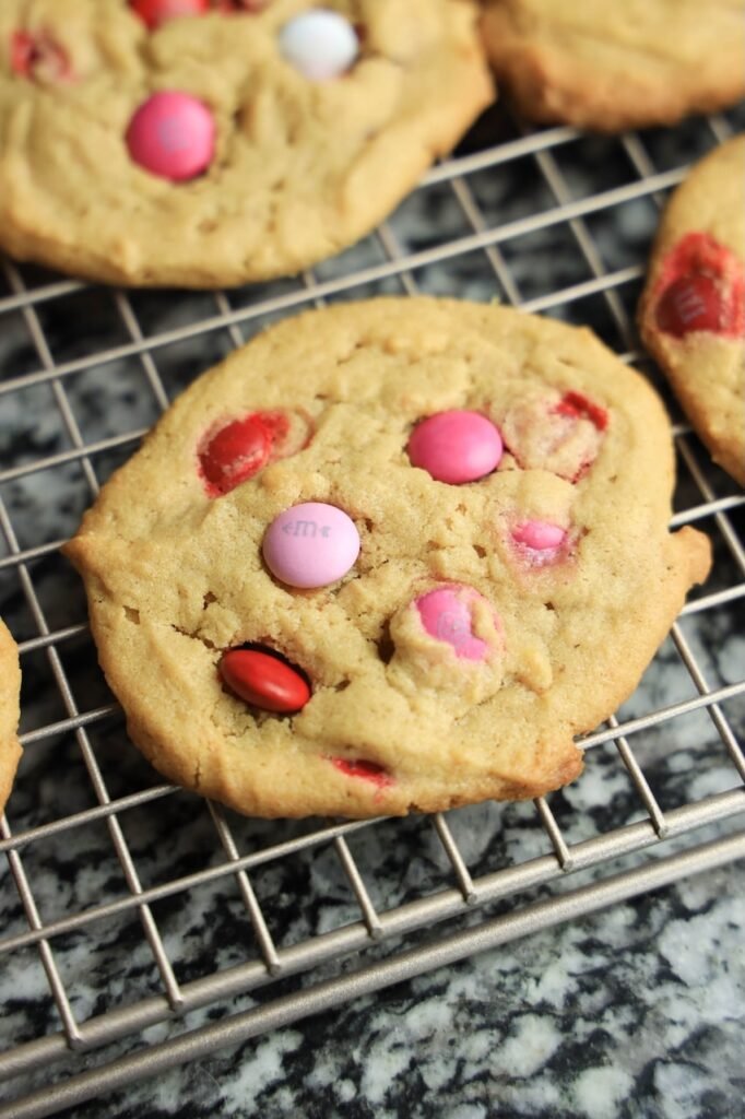 Freshly baked Valentine’s M&M cookies cooling on a wire rack, showing soft peanut butter cookies studded with red, pink, and white M&M candies.