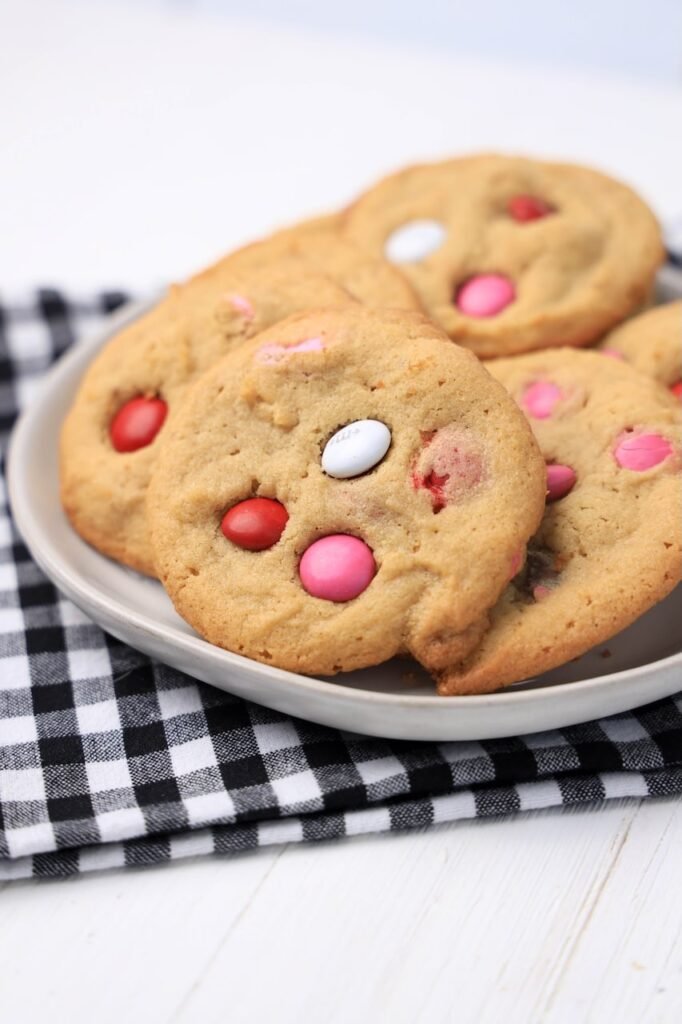 A plate of soft Valentine’s M&M cookies arranged on a black-and-white cloth, showing golden peanut butter cookies with red, pink, and white M&M candies on top.