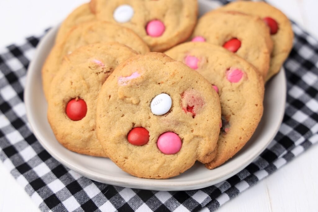 A plate of soft Valentine’s M&M cookies arranged on a black-and-white cloth, showing golden peanut butter cookies dotted with red, pink, and white M&M candies.