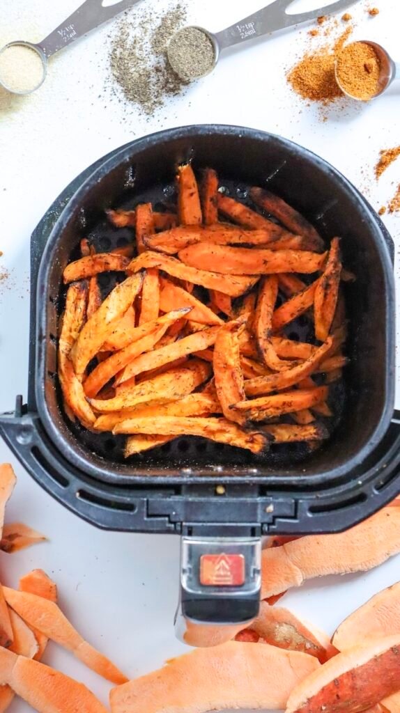 Overhead view of seasoned sweet potato fries arranged in a single layer inside an air fryer basket, lightly browned on the edges as they cook, with loose spices visible around the basket.