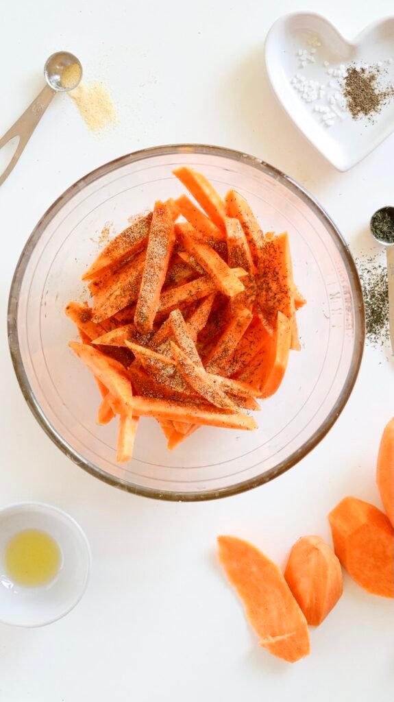 Overhead view of sliced sweet potato fries in a clear glass bowl, lightly coated with olive oil and sprinkled with paprika, garlic powder, dried dill, salt, and black pepper before air frying.