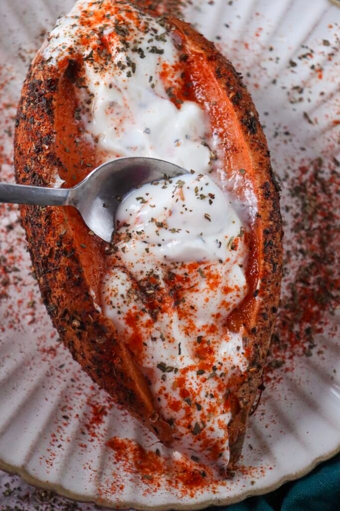 Air fryer sweet potato jacket split open and topped with Greek yogurt and herbs, with a spoon scooping the creamy center.