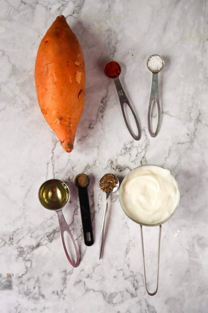 Flat lay of ingredients for air fryer sweet potato jackets with Greek yogurt including one whole sweet potato, olive oil, paprika, salt, pepper, dried basil, and a bowl of plain yogurt on a marble countertop.
