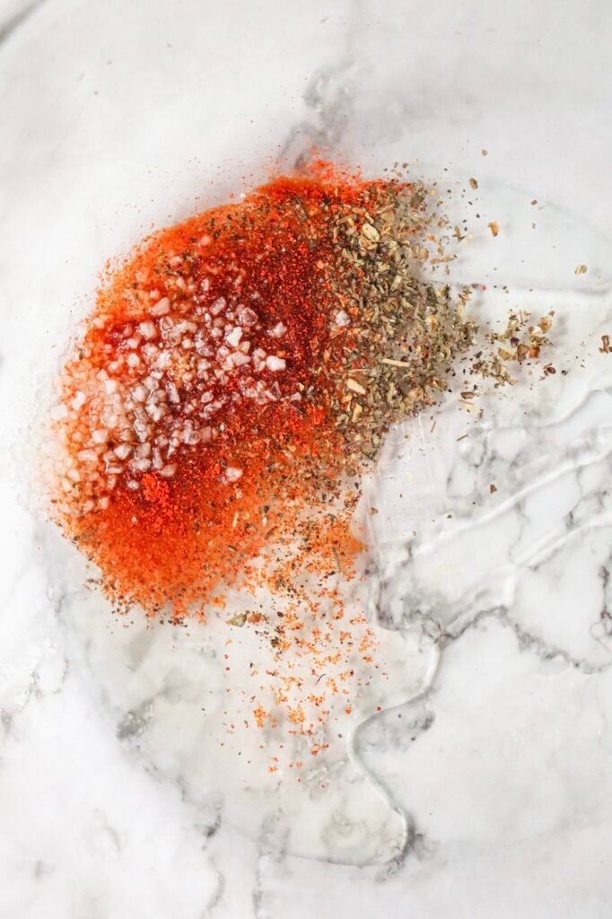 Close up of paprika, coarse salt, dried basil, black pepper, and olive oil on a marble surface for seasoning before cooking.