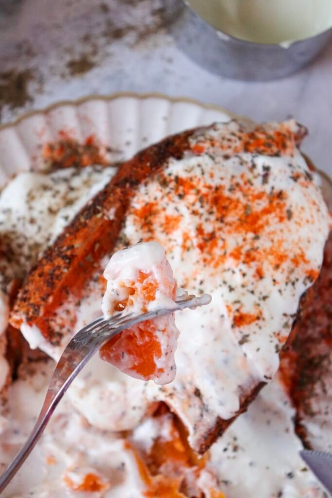 Fork holding a bite of air fryer sweet potato topped with Greek yogurt, showing fluffy orange interior and crispy seasoned skin in the background.