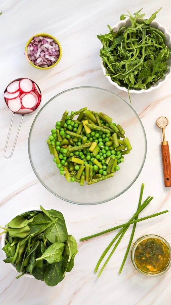 Overhead view of blanched asparagus and peas in a glass bowl surrounded by spinach, arugula, sliced radishes, red onion, chives, and lemon Dijon dressing on a light marble surface.