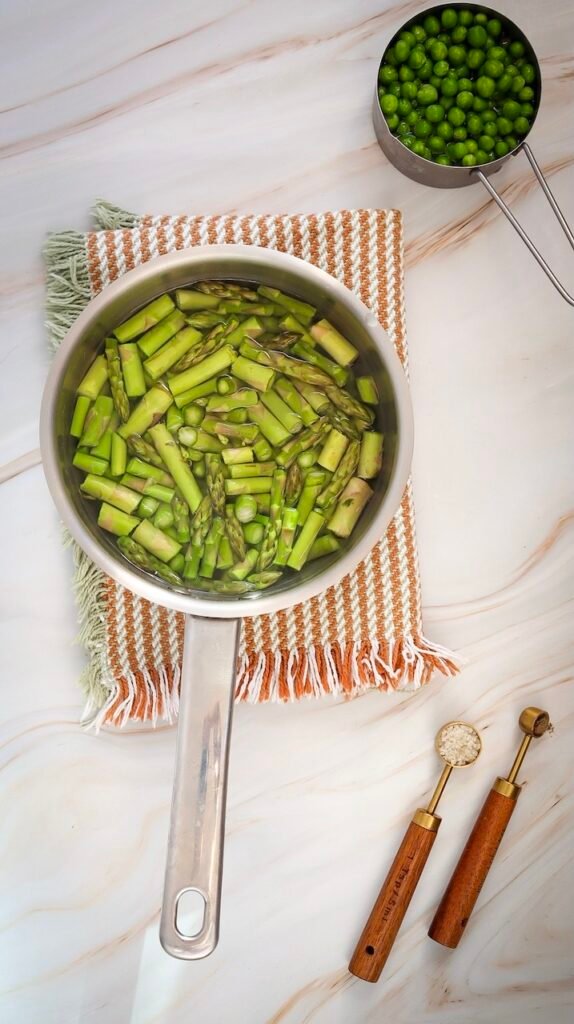 Overhead view of chopped asparagus pieces in a saucepan filled with hot water during blanching, with fresh peas nearby on a light marble surface.