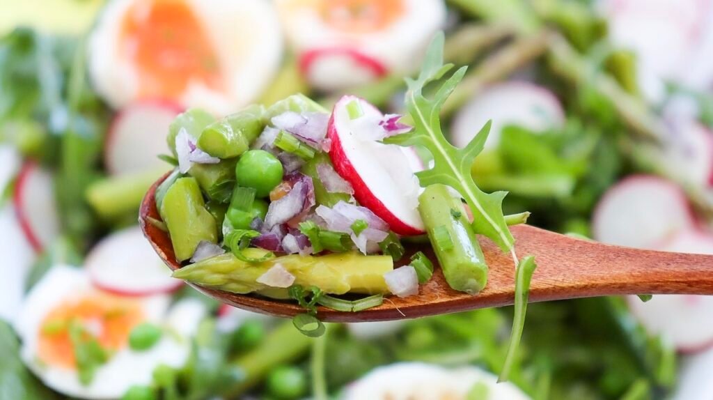 Close-up of a spoonful of asparagus pea salad with peas, radishes, red onion, and greens, with soft-boiled eggs in the background.