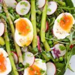 Close-up of asparagus and pea salad topped with halved soft-boiled eggs, sliced radishes, red onion, and fresh greens on a white serving platter.
