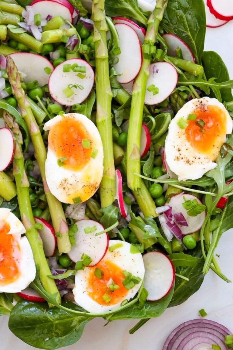 Close-up of asparagus and pea salad topped with halved soft-boiled eggs, sliced radishes, red onion, and fresh greens on a white serving platter.