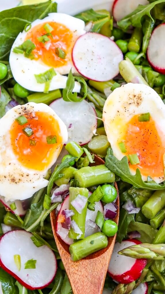 Close-up of asparagus pea salad topped with halved soft-boiled eggs, radishes, red onion, and fresh greens on a serving platter.