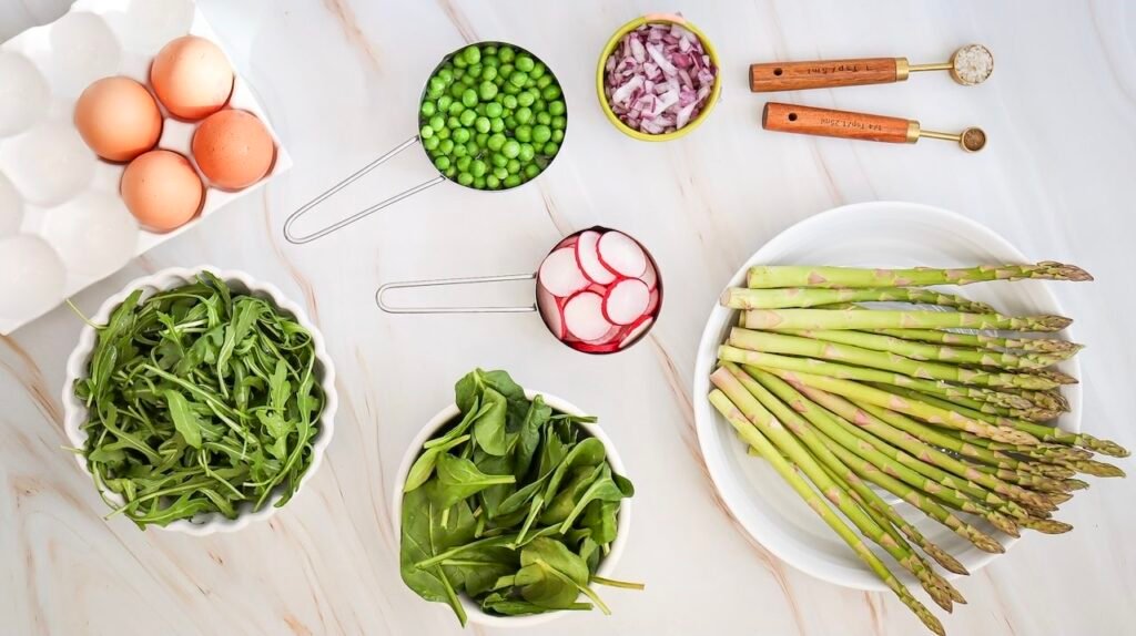 Overhead view of fresh ingredients for asparagus and pea salad including whole asparagus spears, peas, sliced radishes, red onion, spinach, arugula, and eggs arranged on a light marble surface.