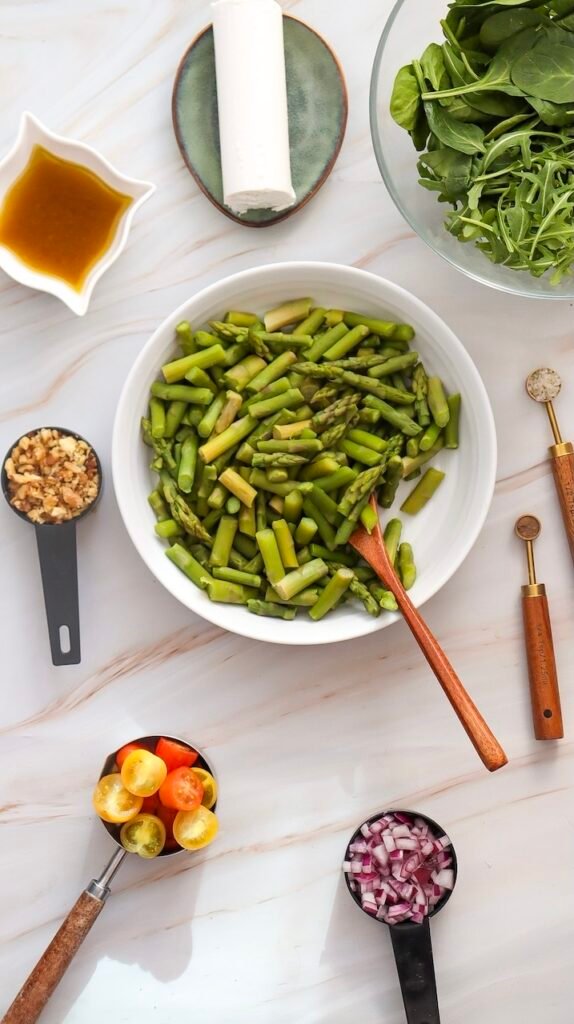 Overhead view of blanched asparagus in a white bowl with a wooden spoon, surrounded by salad ingredients including greens, goat cheese, walnuts, cherry tomatoes, and red onion.