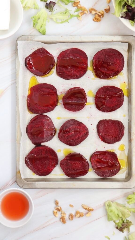 Thin slices of deep red beetroot arranged on a parchment-lined baking sheet, drizzled with olive oil and ready to be roasted, with salad greens and walnuts visible around the tray on a light marble surface.