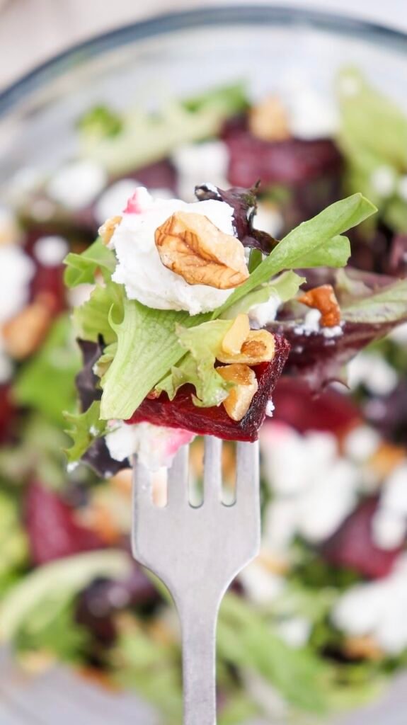 Fork holding a bite of beet goat cheese salad with roasted beet pieces, crumbled goat cheese, chopped walnuts, and mixed greens, with the finished salad softly blurred in the background.