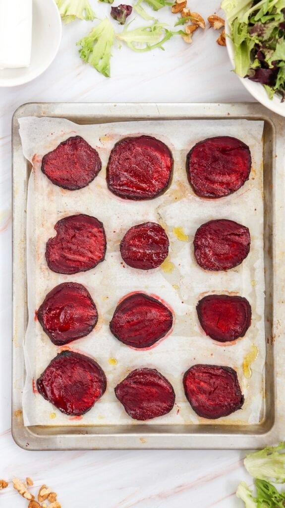 Roasted beetroot slices arranged on a parchment-lined baking sheet, tender and lightly caramelized, ready to be added, with scattered salad greens and chopped walnuts visible around the pan on a light marble surface.
