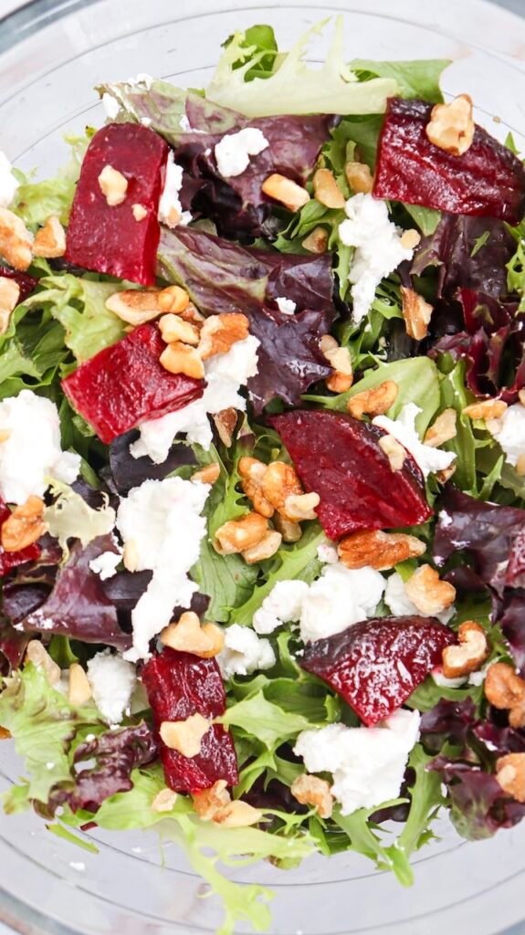 Close-up view of beet goat cheese salad made with mixed salad greens, roasted beet pieces, crumbled goat cheese, and chopped walnuts in a clear glass bowl, showing the finished salad before adding dressing.