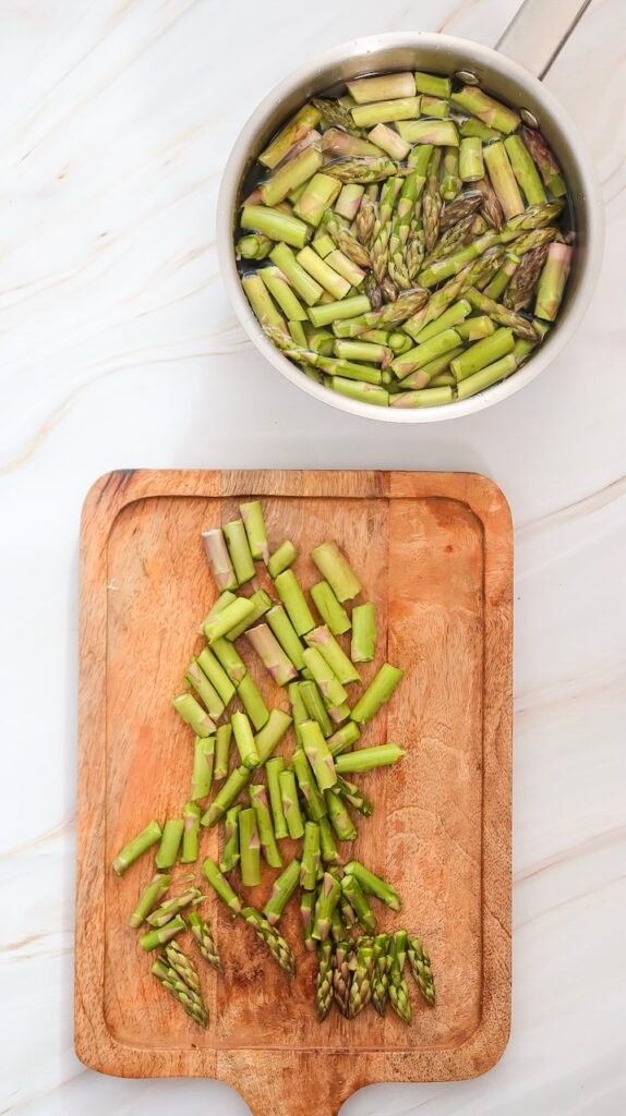 Overhead view of chopped asparagus on a wooden cutting board with asparagus blanching in a pot of water, showing the preparation step for goat cheese and asparagus salad.