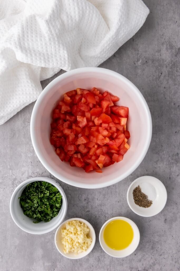 Overhead view of a bowl of diced tomatoes surrounded by chopped basil, minced garlic, olive oil, salt, and black pepper on a gray surface, showing the ingredients prepared for mixing classic italian topping.
