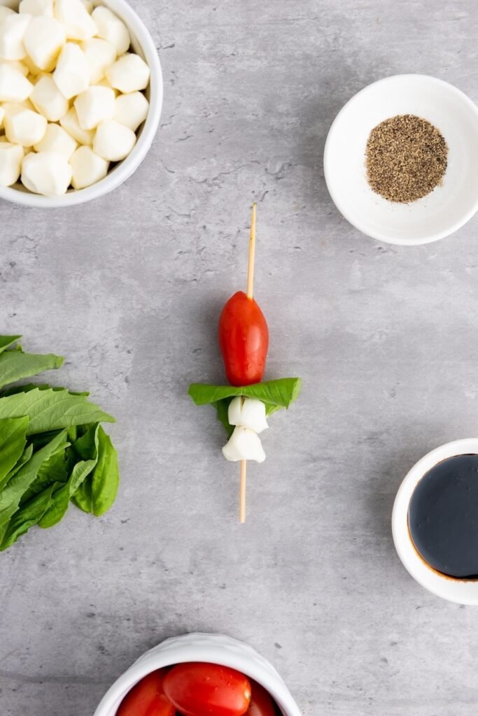 Overhead view of a wooden skewer with mozzarella pearls, a folded basil leaf, and a cherry tomato threaded on in order, surrounded by ingredients, demonstrating the proper layering step for assembling.