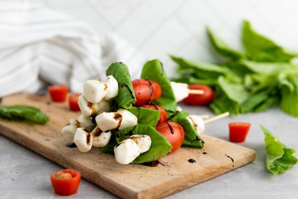 Caprese skewers stacked on a wooden board with mozzarella pearls, basil leaves, and cherry tomatoes drizzled with balsamic glaze, shown as a finished easy party appetizer ready to serve.