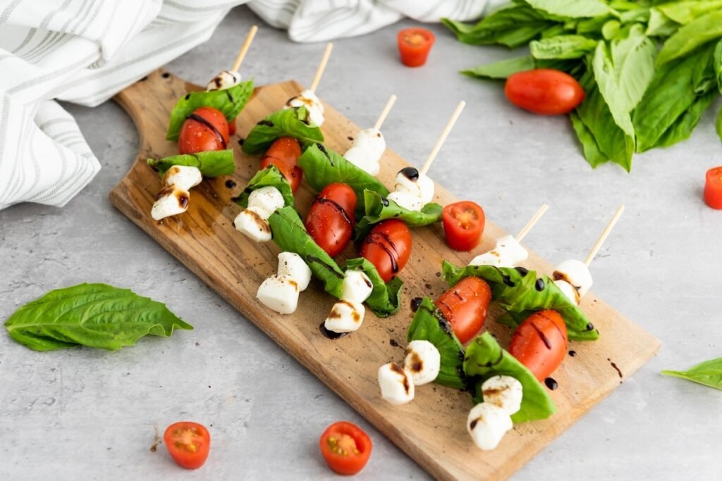 Caprese skewers arranged on a wooden board with mozzarella, basil, and cherry tomatoes, drizzled lightly with balsamic glaze and sprinkled with black pepper, ready to serve as a fresh appetizer.