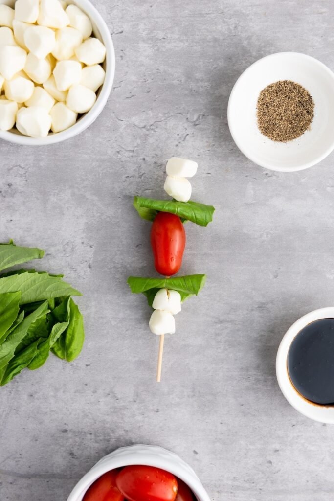 Overhead view of a fully assembled appetizer with mozzarella pearls, basil leaves, and a cherry tomato stacked on a wooden skewer, surrounded by ingredients, showing the completed assembly step before adding balsamic glaze.