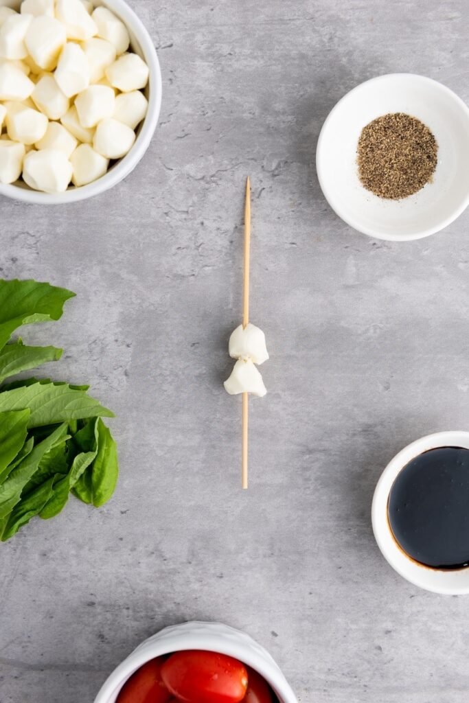 Overhead view of a wooden skewer with two mozzarella pearls threaded on it, surrounded by basil leaves, cherry tomatoes, balsamic glaze, and black pepper, showing the first step in assembling caprese skewers.