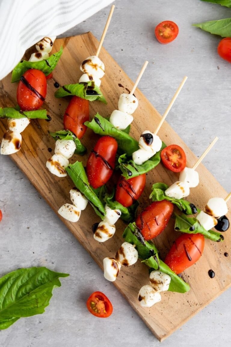 Caprese skewers arranged diagonally on a wooden board with mozzarella, basil, and cherry tomatoes, lightly drizzled with balsamic glaze and sprinkled with black pepper, shown as a finished party-style appetizer.