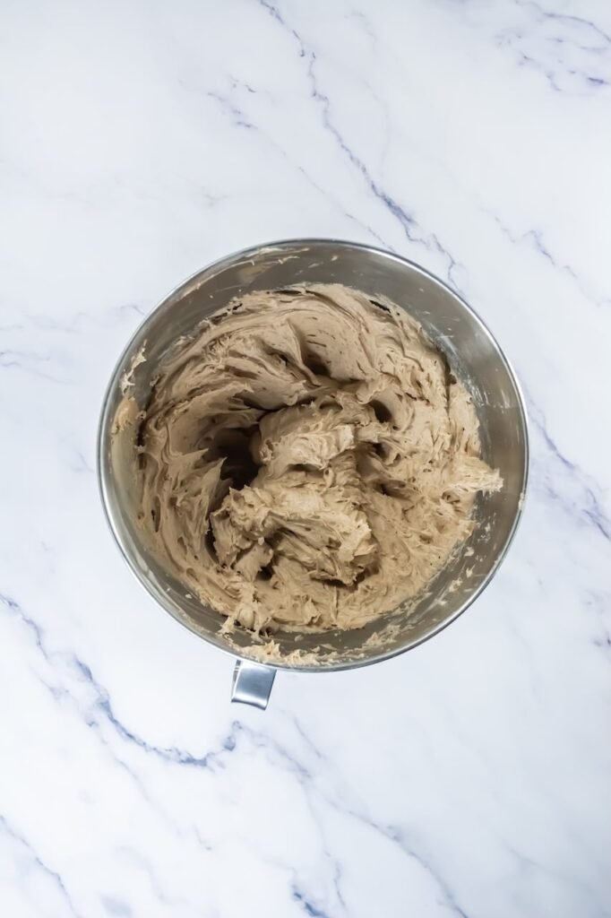 Overhead view of whipped cinnamon buttercream frosting in a stand mixer bowl, showing smooth, fluffy texture ready to pipe.