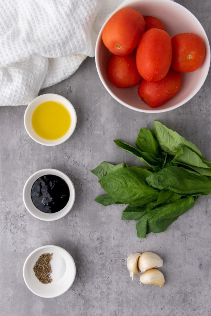 Overhead view of fresh bruschetta ingredients arranged on a gray surface, including a bowl of Roma tomatoes, fresh basil leaves, garlic cloves, olive oil, balsamic glaze, salt, and black pepper, ready to prepare classic bruschetta.