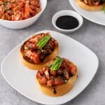 Two slices of toasted bread topped with fresh tomato basil bruschetta and drizzled with balsamic glaze on a white plate, with extra tomato topping and glaze in the background, ready to serve as an appetizer.