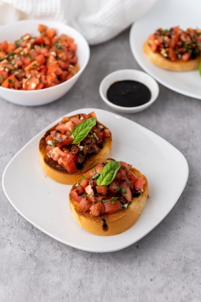 Two slices of toasted bread topped with fresh tomato basil bruschetta and drizzled with balsamic glaze on a white plate, with extra tomato topping and glaze in the background, ready to serve as an appetizer.