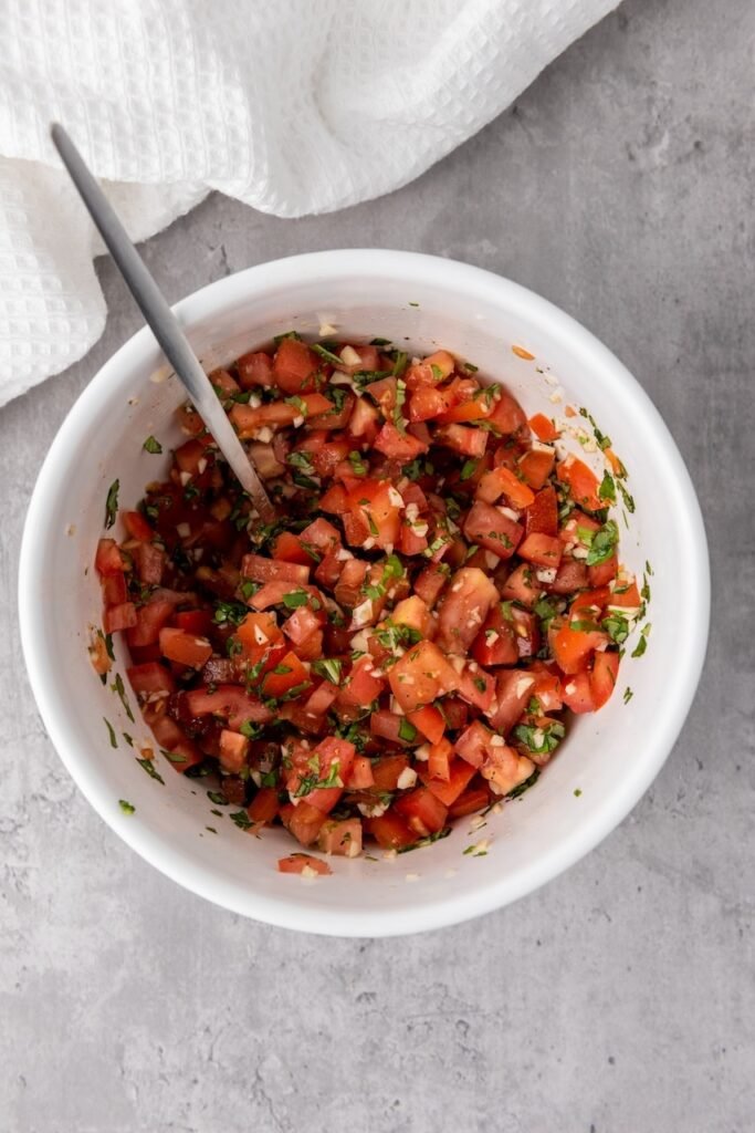 Overhead view of a bowl filled with mixed topping made from diced tomatoes, chopped basil, minced garlic, olive oil, and balsamic glaze, with a spoon resting inside, ready to serve over toasted bread.