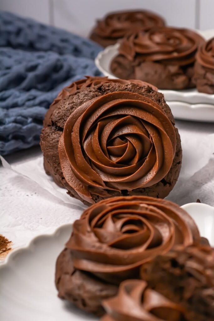 Close-up view of a thick chocolate cake cookie topped with a large swirl of smooth chocolate frosting, with additional frosted cookies softly blurred in the background.