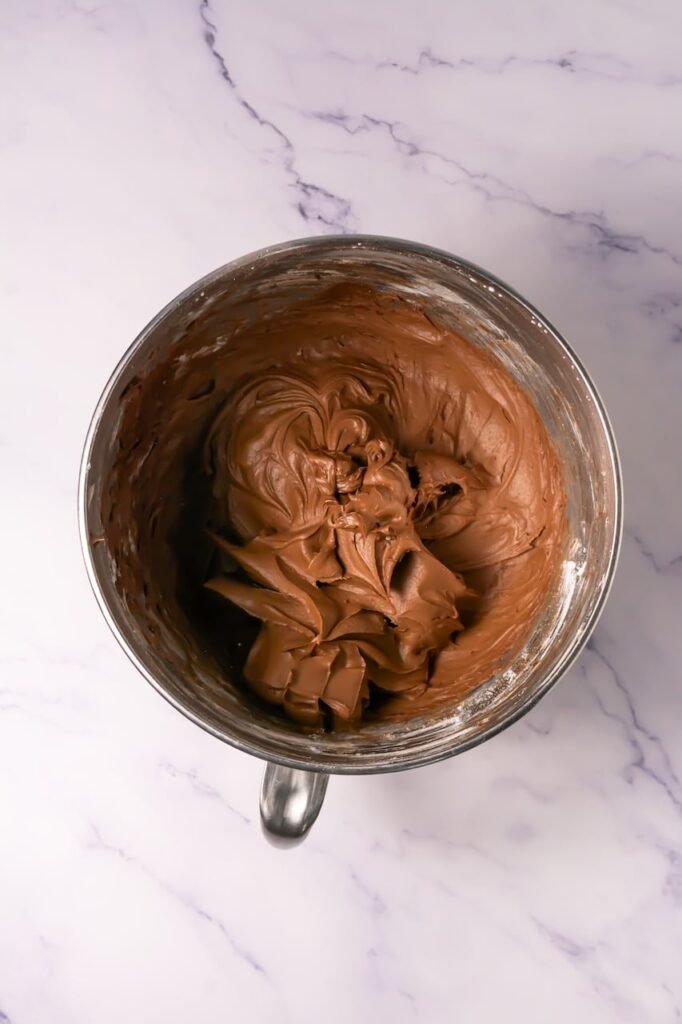 Overhead view of a stand mixer bowl filled with smooth, whipped chocolate cream cheese frosting being mixed on a white marble countertop.
