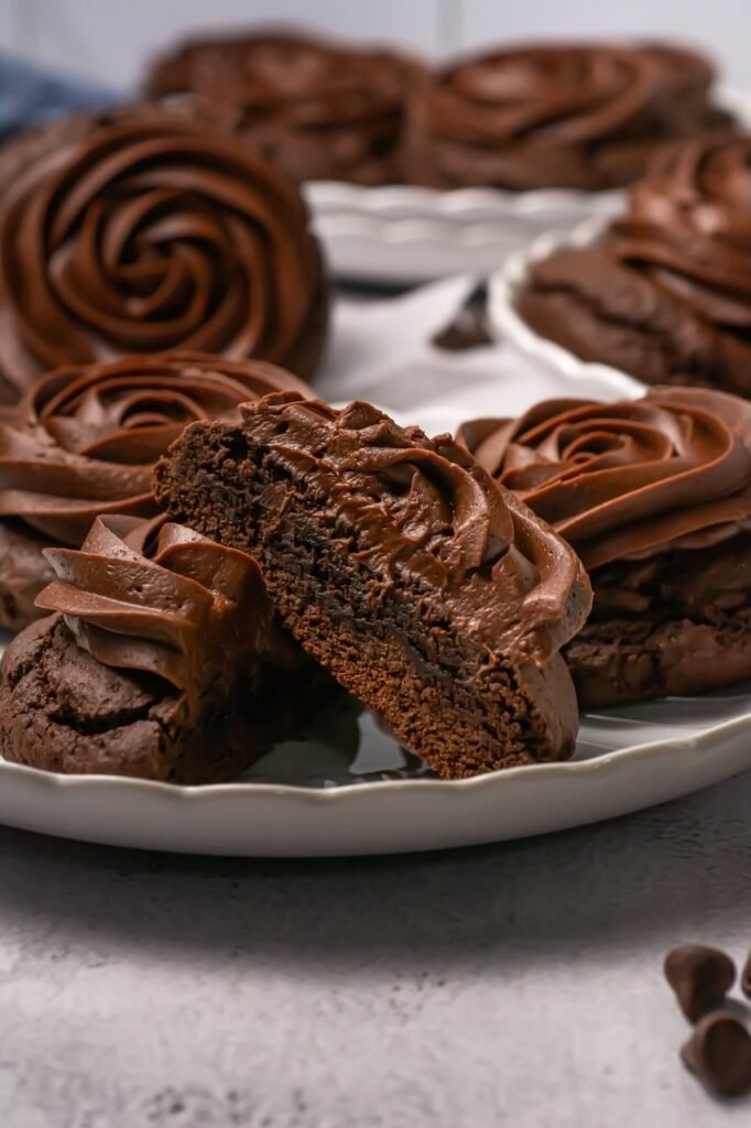 Close-up of a thick chocolate cake cookie cut in half to show its soft, cake-like center and rich chocolate frosting swirl on top, with additional frosted cookies in the background.