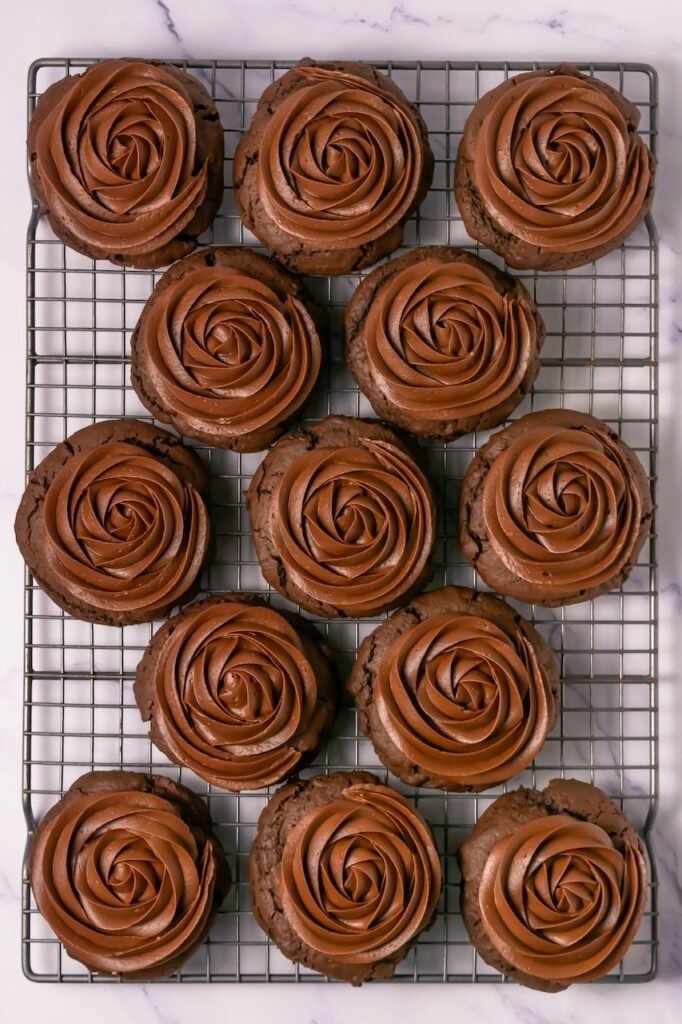 Overhead view of thick chocolate cake cookies topped with large swirls of smooth chocolate frosting arranged on a cooling rack, showing bakery-style texture and rich chocolate color.