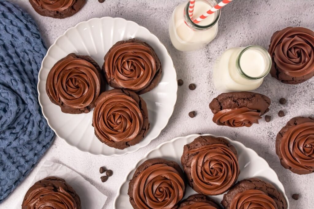 Overhead view of thick chocolate cake cookies topped with large swirls of chocolate frosting arranged on white plates with small glass bottles of milk and chocolate chips nearby.