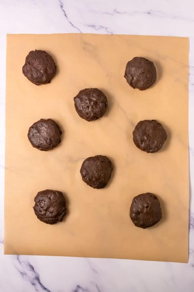 Overhead view of large scoops of chocolate cookie dough spaced evenly on parchment paper, ready to bake into thick desserts.