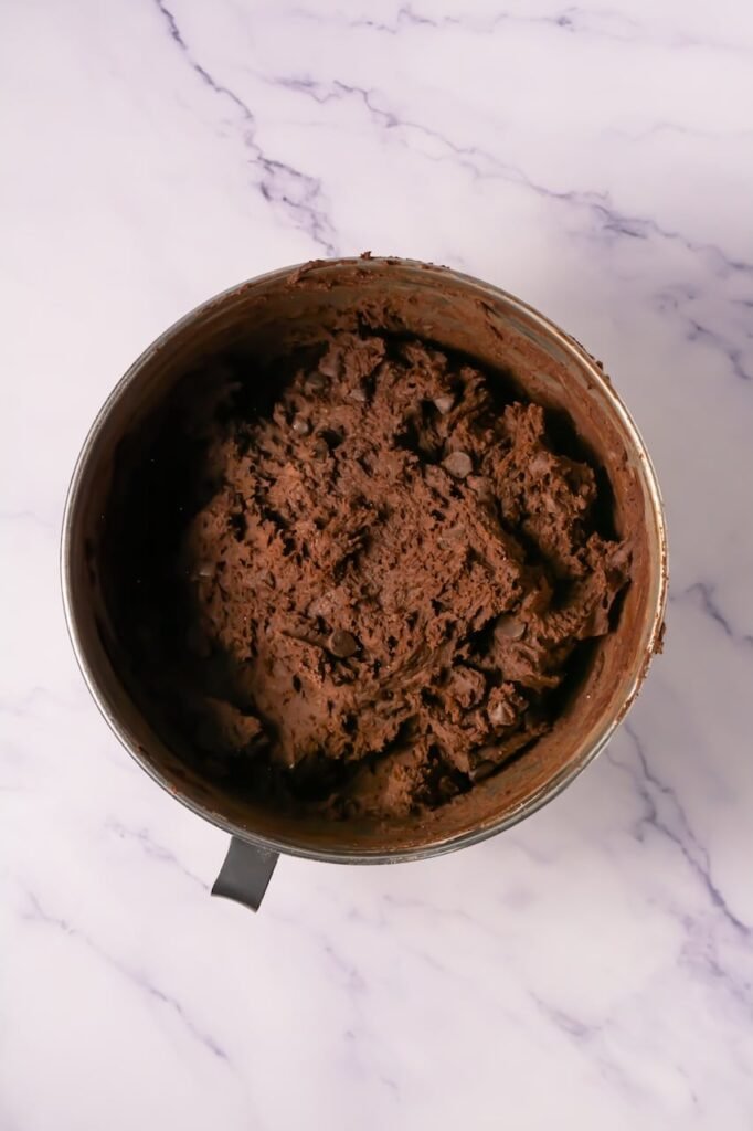 Overhead view of thick chocolate cookie dough mixed with chocolate chips inside a stand mixer bowl, showing the proper texture before scooping for Crumbl-style chocolate cake cookies.