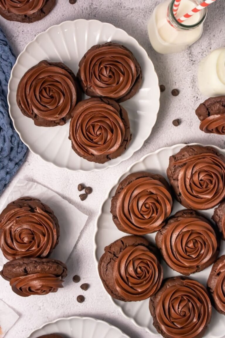 Overhead view of thick, soft chocolate cake cookies topped with large swirls of smooth chocolate frosting arranged on white plates with small bottles of milk nearby.