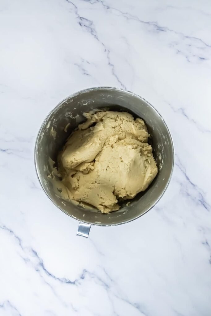 Overhead view of thick, fully mixed Crumbl churro cookie dough inside a stand mixer bowl on a white marble surface, showing the smooth but sturdy texture before scooping.