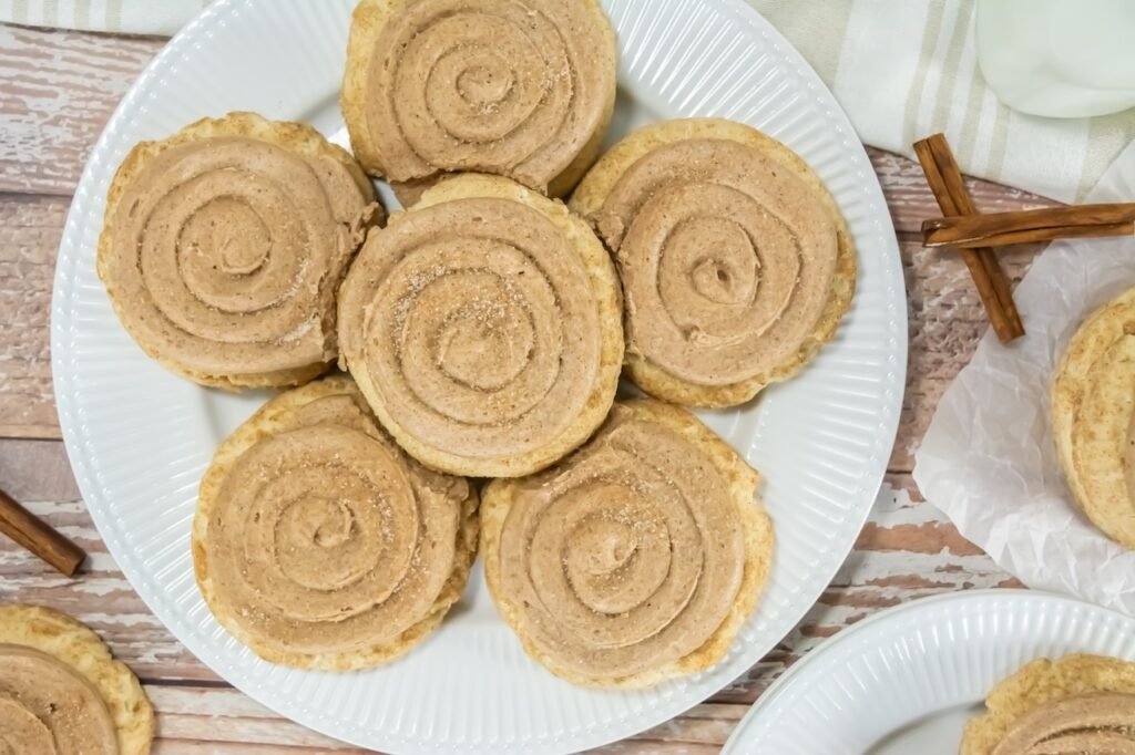 Overhead view of Crumbl churro cookies arranged on a white plate, each topped with a spiral of cinnamon buttercream and lightly dusted with cinnamon sugar, with cinnamon sticks on the side.
